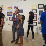 Young girl points out something in the distance to her father as people walk around in the background looking at colorful youth art on the walls